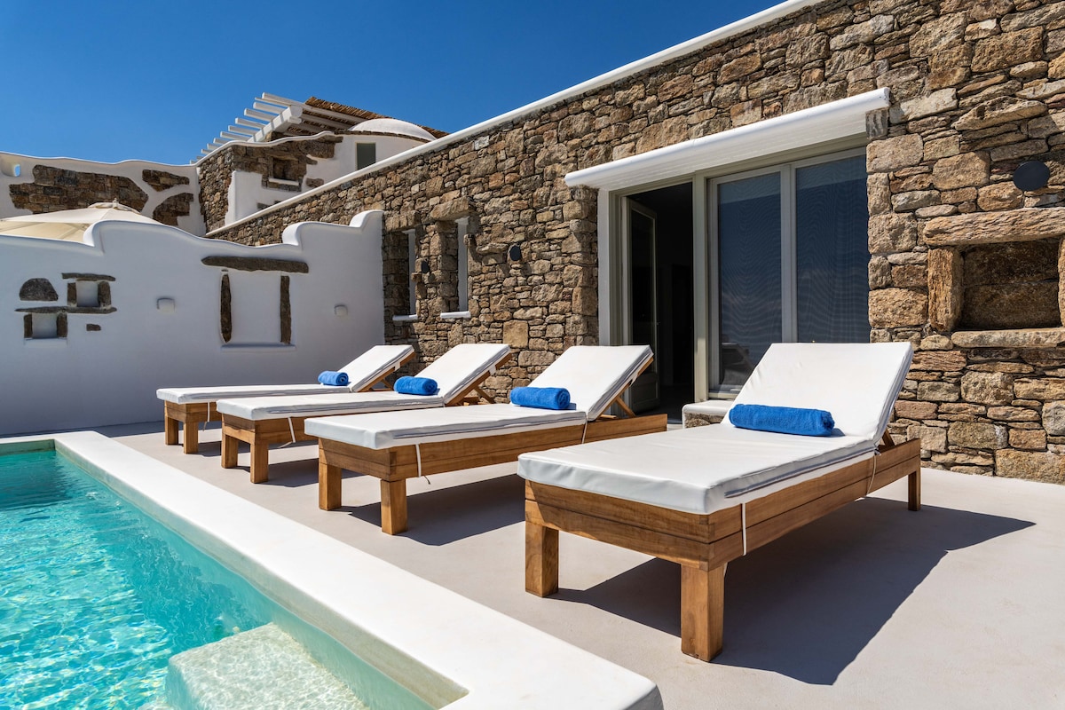 Four loungers with plush white cushions and blue towels are arranged near a private infinity pool. The stone villa features large glass doors, allowing sunlight to stream into the interior. The surrounding area displays traditional architecture, complemented by a clear blue sky.