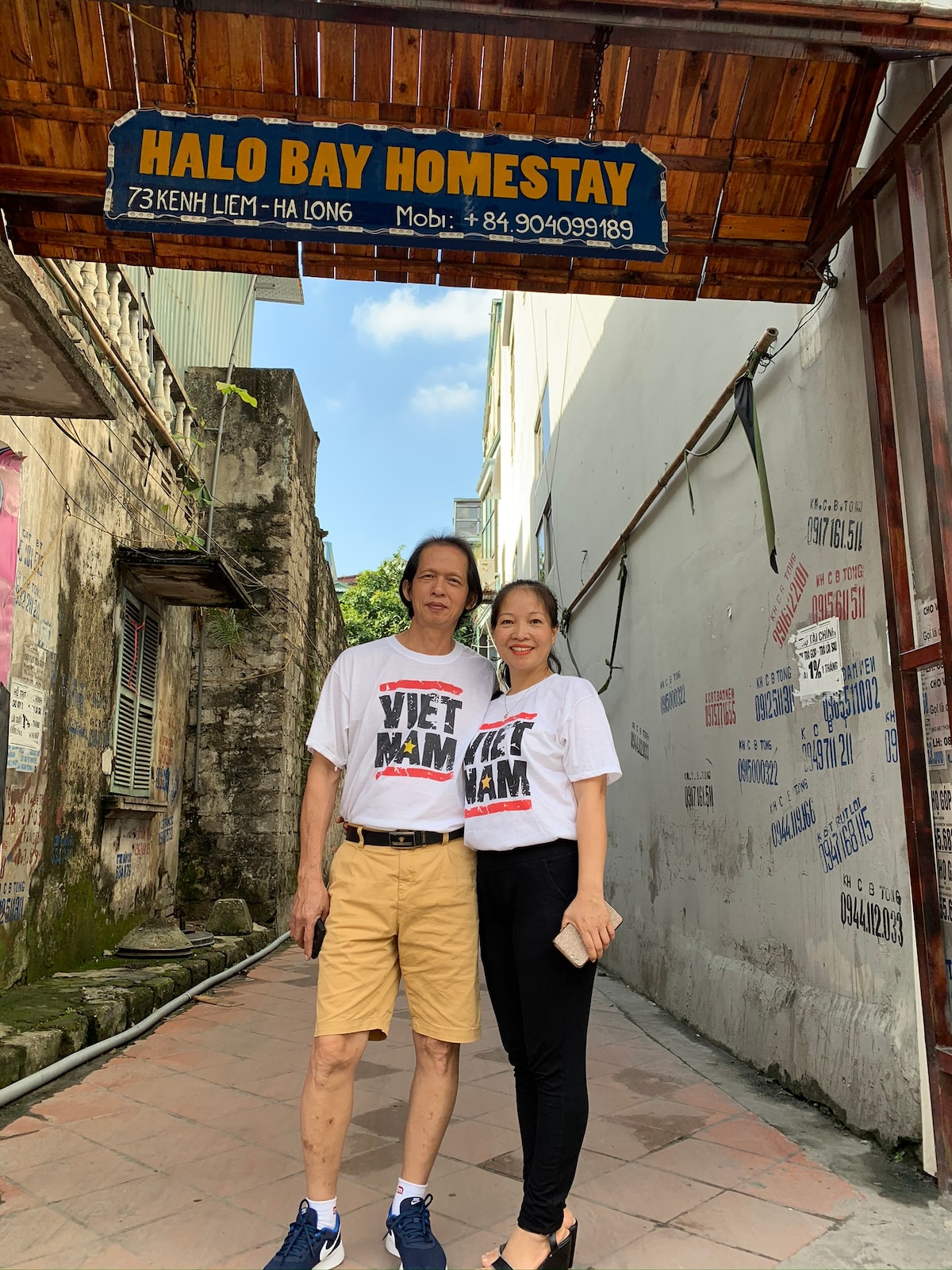 The entrance to Halo Bay Homestay is marked by a wooden sign hanging above, displaying the property name. A couple stands together in front, wearing matching T-shirts, with a street visible behind them showcasing an urban setting with brick walls and nearby buildings.