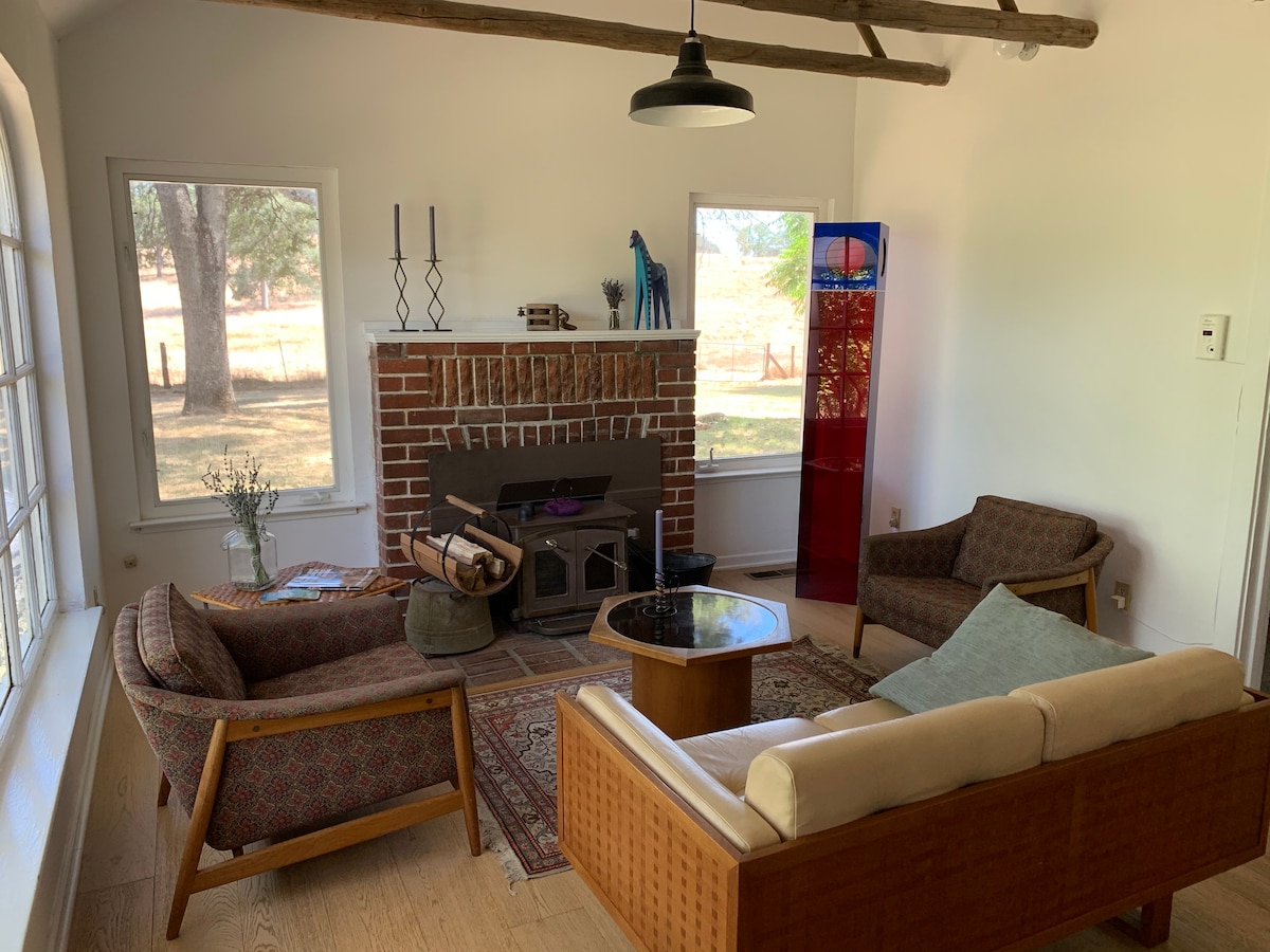 The living area exhibits wooden beams overhead and a stylish fireplace. Two armchairs and a sofa are arranged around a wooden coffee table on a patterned rug. Large windows allow natural light, framing views of the surrounding landscape.