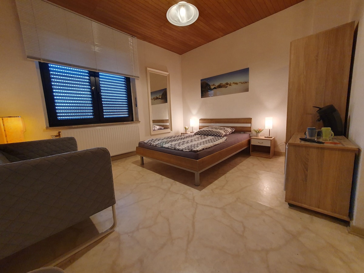 A cozy bedroom features a spacious bed with patterned bedding, situated next to a bedside table holding a kettle and two cups. A comfortable armchair is visible, along with a mirror reflecting natural light from the window. Soft lighting is provided by a nearby lamp.