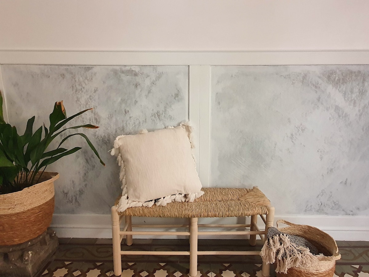 A natural wood bench is positioned against a light gray wall, accompanied by a textured white cushion. A potted plant with broad leaves is placed to one side, and woven baskets are arranged on the floor, adding a touch of organic texture.