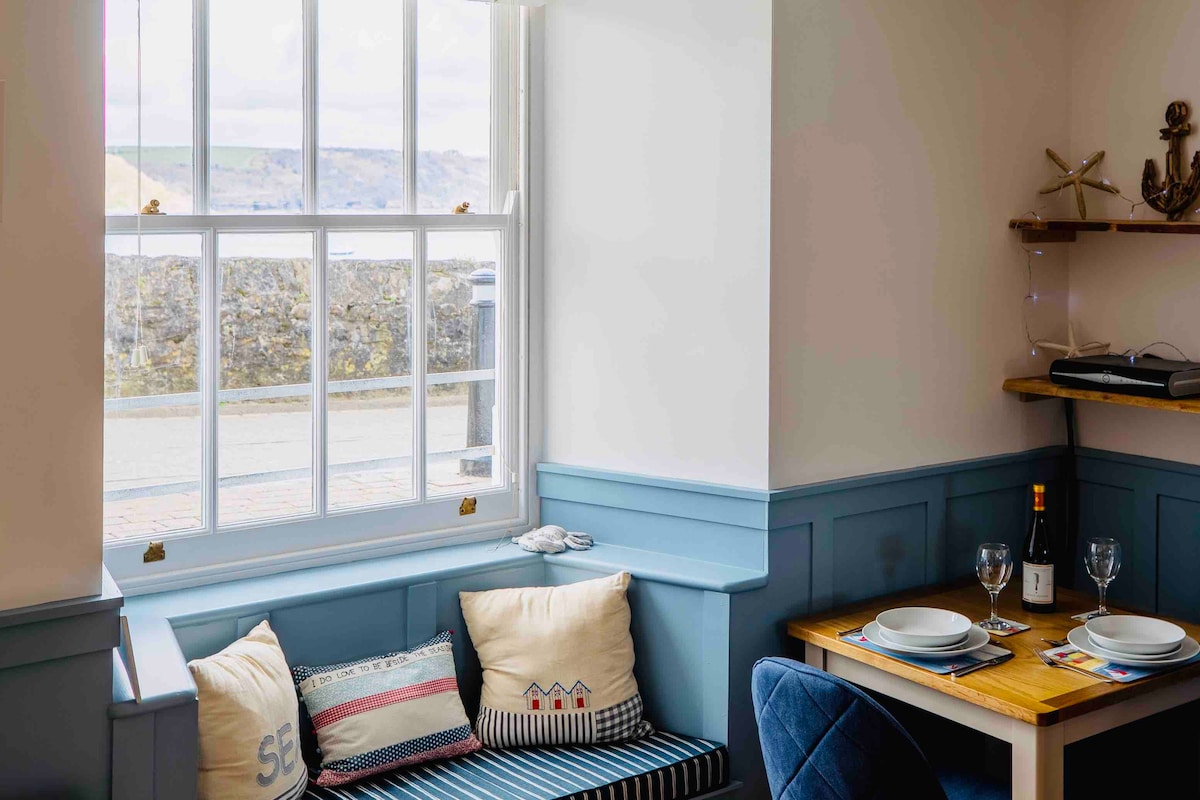 A cozy seating area is illuminated by natural light from a large window, providing views of the surroundings. A neatly set dining table with plates and glassware is visible, alongside decorative pillows on the built-in bench, adding comfort to the space.