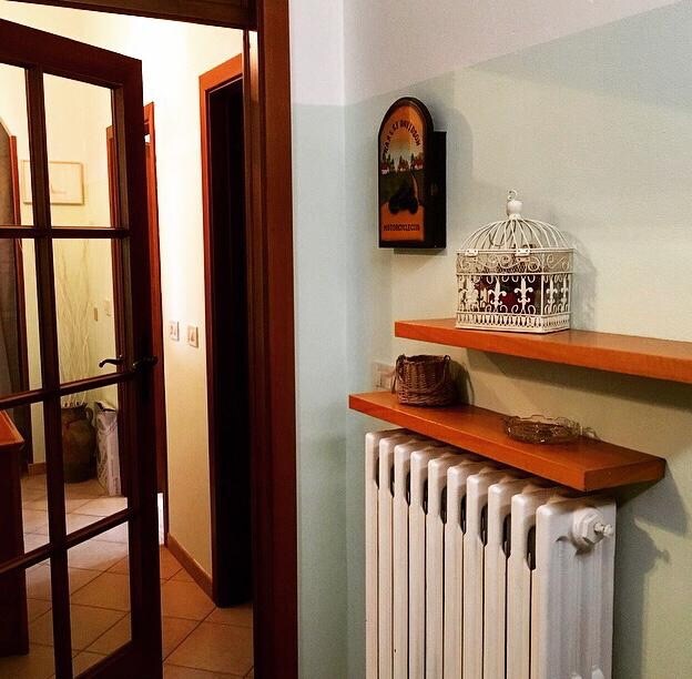 An entryway is illustrated with a wooden shelf displaying a decorative birdcage and small baskets. A white radiator is positioned beneath the shelf. Light beige and soft green walls create a welcoming atmosphere. Glass-paned doors lead to other areas of the space.