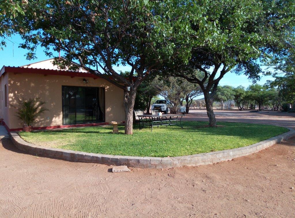 A simple farmhouse is set in a grassy area, surrounded by trees providing shade. A patio with a table and chairs is visible in front of the house, inviting relaxation. The landscape consists of red earth and sparse vegetation, enhancing the natural surroundings.