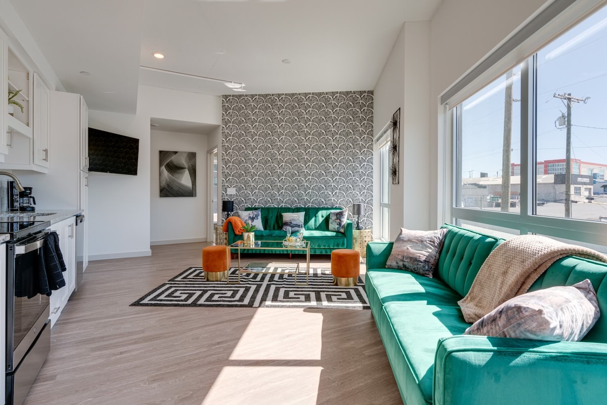 A modern living area features green sofas and an accent rug with geometric patterns. Large windows allow natural light to fill the space, highlighting the sleek cabinetry and decor. A stylish coffee table is centrally located, enhancing the functional and inviting atmosphere.