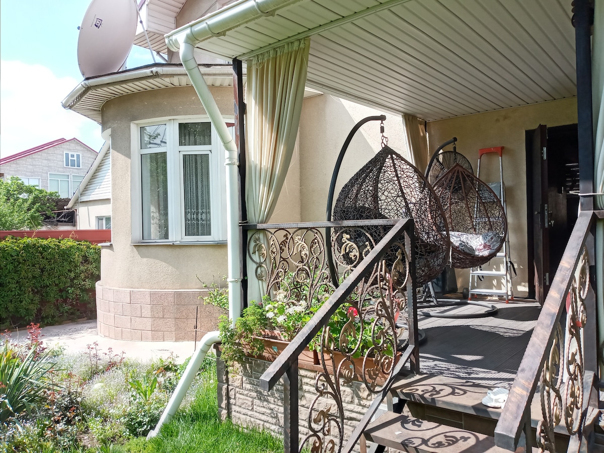 A welcoming entrance features two hanging chairs, offering a relaxing spot on a covered porch. Lush greenery surrounds the area, with a well-maintained pathway leading toward the home. The structure exhibits a blend of natural light and shaded areas, enhancing the inviting atmosphere.