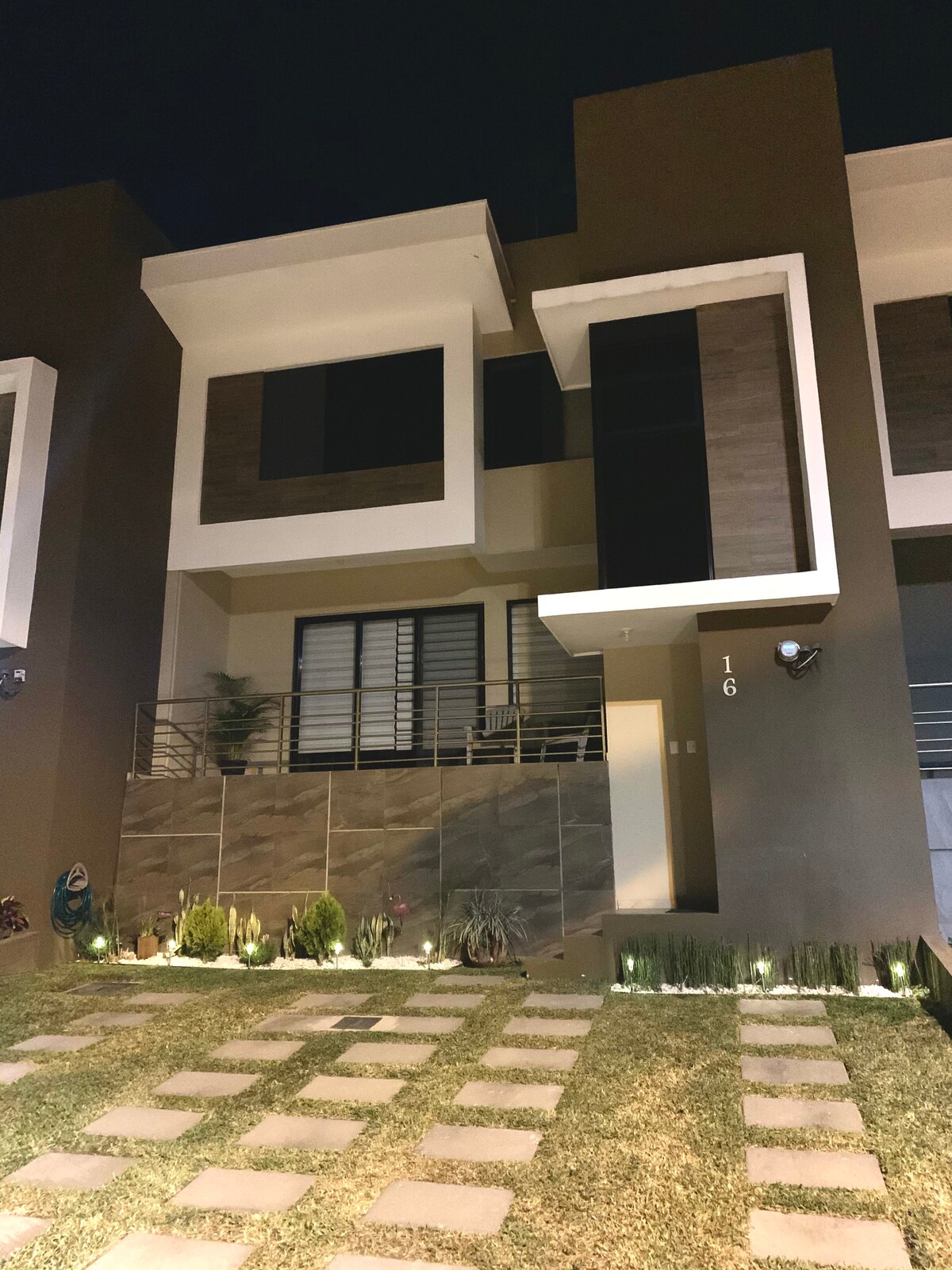 The townhouse exterior is showcased at night, featuring a modern façade with large windows and a subtle light accent. A well-manicured lawn with stepping stones leads to the entrance. Decorative plants and soft ground lighting enhance the inviting appearance.