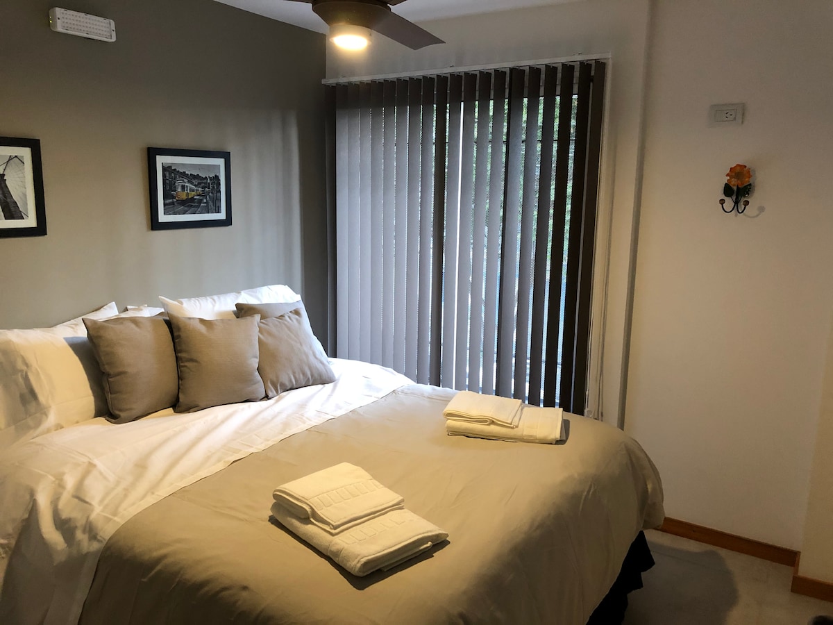 A spacious bedroom features a large bed adorned with neutral bedding and decorative pillows. Fresh towels are neatly arranged at the foot of the bed. Natural light filters through the vertical blinds, illuminating the room's calming color palette.