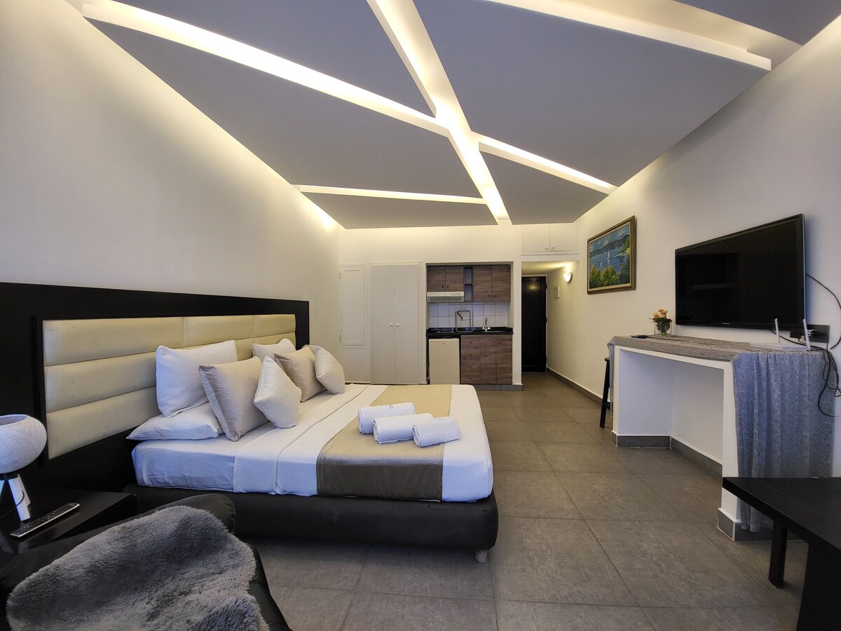 A cozy studio interior features a spacious bed with neutral bedding and decorative cushions. A kitchenette is visible in the background, with light wood cabinetry and modern appliances. Soft overhead lighting creates an inviting atmosphere, complemented by a flat-screen TV mounted on the wall.