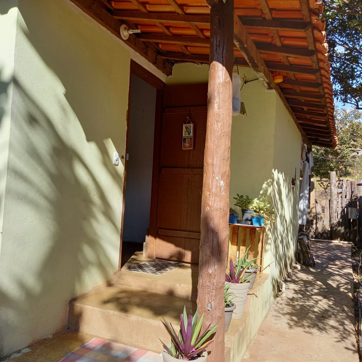 An entrance is framed by a wooden post and features a simple door adorned with a decorative sign. Potted plants are arranged beside the entryway, while warm sunlight casts gentle shadows on the exterior walls, highlighting the rustic charm of the structure.