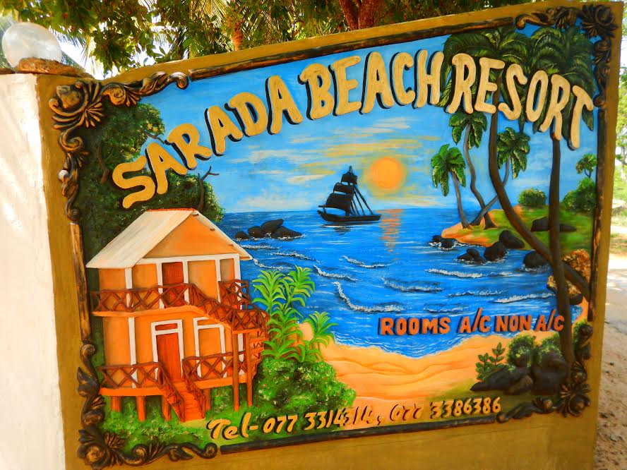 The colorful sign for Sarada Beach Resort features a seaside scene with a sailboat, tropical trees, and a sandy beach. A two-story building is depicted, highlighting available rooms. The contact information is displayed at the bottom, ensuring easy access for inquiries.