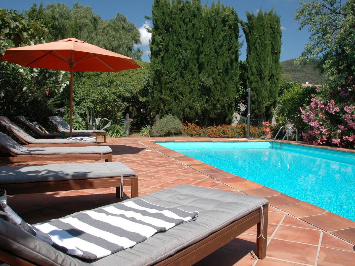 A tranquil outdoor area features a blue swimming pool surrounded by lush greenery and vibrant flowers. Several lounge chairs with striped towels are positioned nearby, and a large orange umbrella provides shade. The sunlit scene captures a serene atmosphere perfect for relaxation.