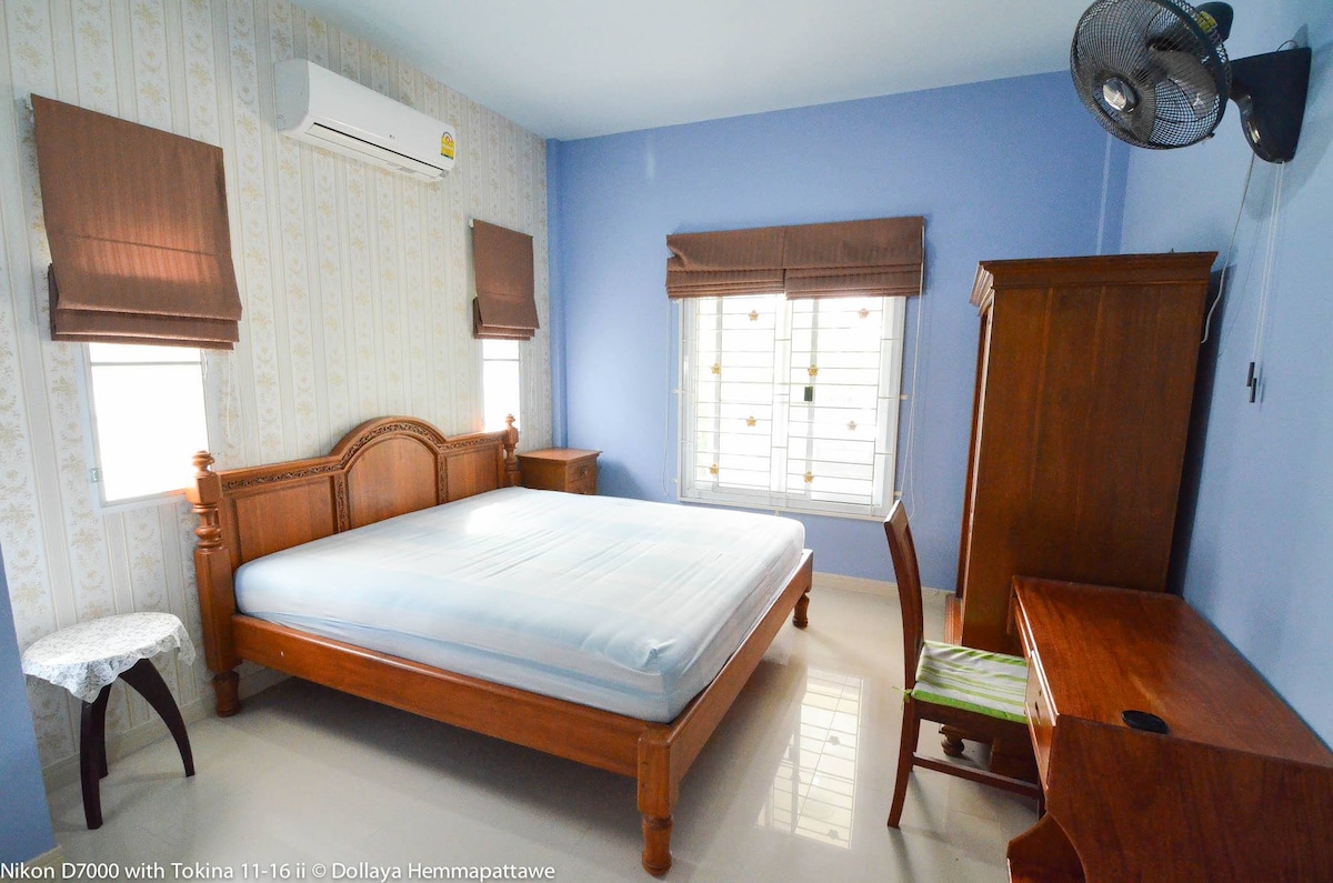 A spacious bedroom is furnished with a wooden bed frame that accommodates a large mattress. A desk and chair sit against the wall, alongside a wardrobe. Natural light filters through the window, which is dressed with brown curtains, set against a cool blue wall.