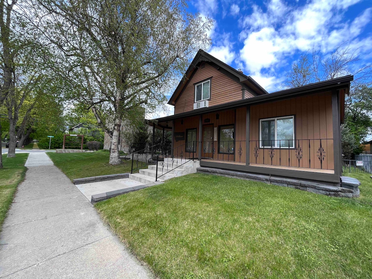 The exterior of a two-story home is shown, featuring warm wood tones and large windows. A covered front porch is accessible by stairs, framed by a well-maintained lawn. A concrete pathway leads to the entrance, with trees and greenery visible in the surrounding area.