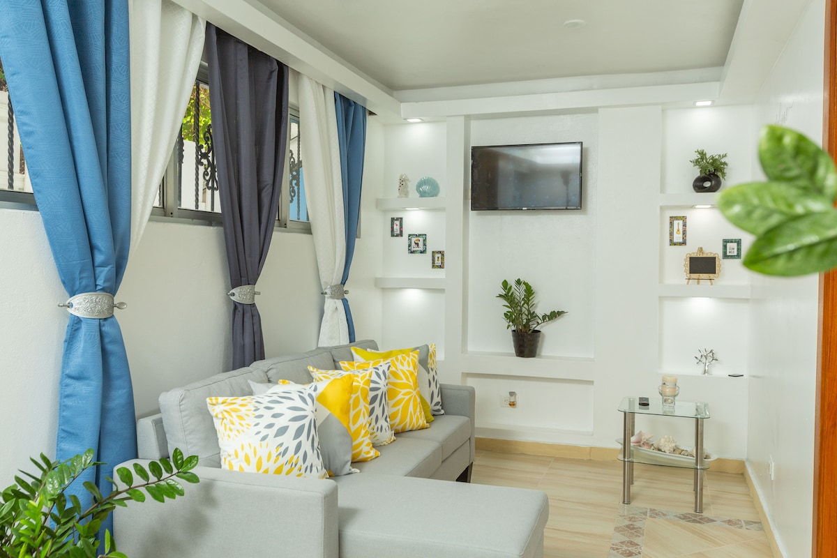 A well-lit living area features a comfortable gray sofa adorned with yellow and blue decorative pillows. Shelves with various decorative items and a small plant are positioned against the bright wall, complemented by a mounted TV. Light curtains frame the windows, allowing natural light to enter.