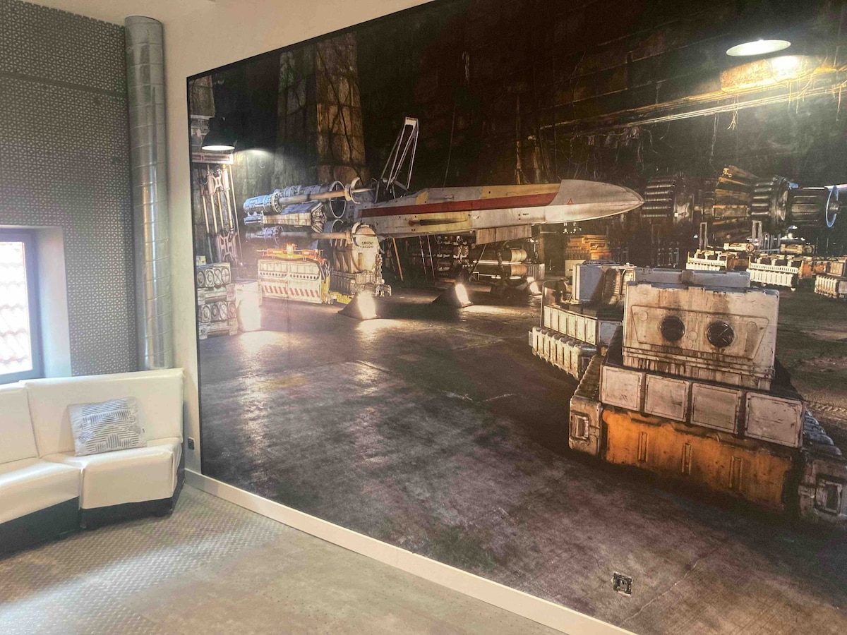 An expansive mural depicts a starship within a hangar setting, accented by industrial elements and ambient lighting. A white seating area is positioned nearby, offering a modern touch against the backdrop of the artwork.