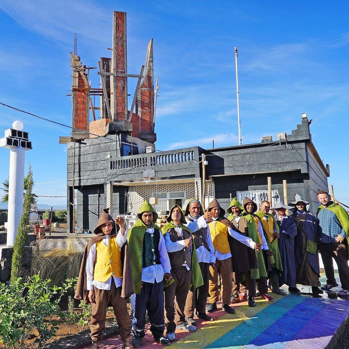 A group of individuals is gathered in front of a reconstructed Orthanc Tower, showcasing a combination of modern architecture and artistic detail. The vibrant, colorful path leading to the structure enhances the appeal of this unique location surrounded by a rural landscape.