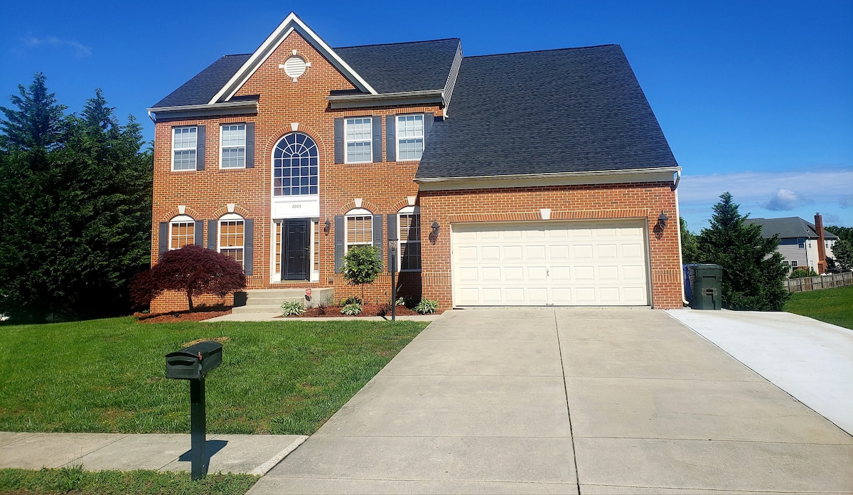 A spacious brick house is set against a clear blue sky, featuring multiple windows and a large front entrance. A driveway leads to a two-car garage, with a green mailbox positioned at the edge of the front yard, which is well-maintained with decorative shrubs.