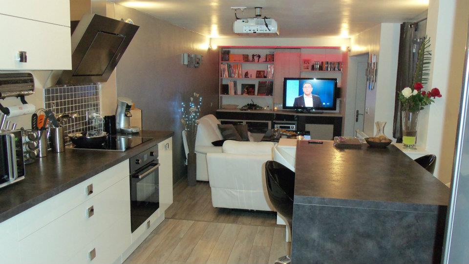 An open living space displays a modern kitchen equipped with sleek cabinetry and appliances, including a coffee machine and oven. A comfortable sofa faces a large TV, and soft lighting creates a relaxed ambiance. A dining table, with stylish chairs, complements the overall design.