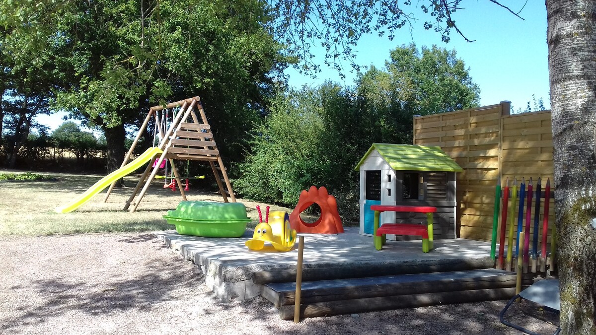 A designated play area is shown, featuring a wooden swing set with a slide, alongside bright plastic play equipment in vibrant colors. A small playhouse with a green roof is included, as well as a picnic table and seating for children, set against a backdrop of greenery.