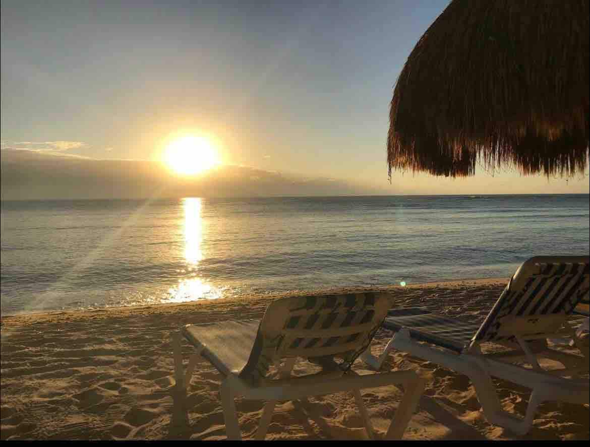 Two lounge chairs are positioned on sandy beach, facing the ocean. The sun sets on the horizon, casting a warm glow over the water's surface. A thatched umbrella partially shades one of the chairs, creating a serene beach setting.