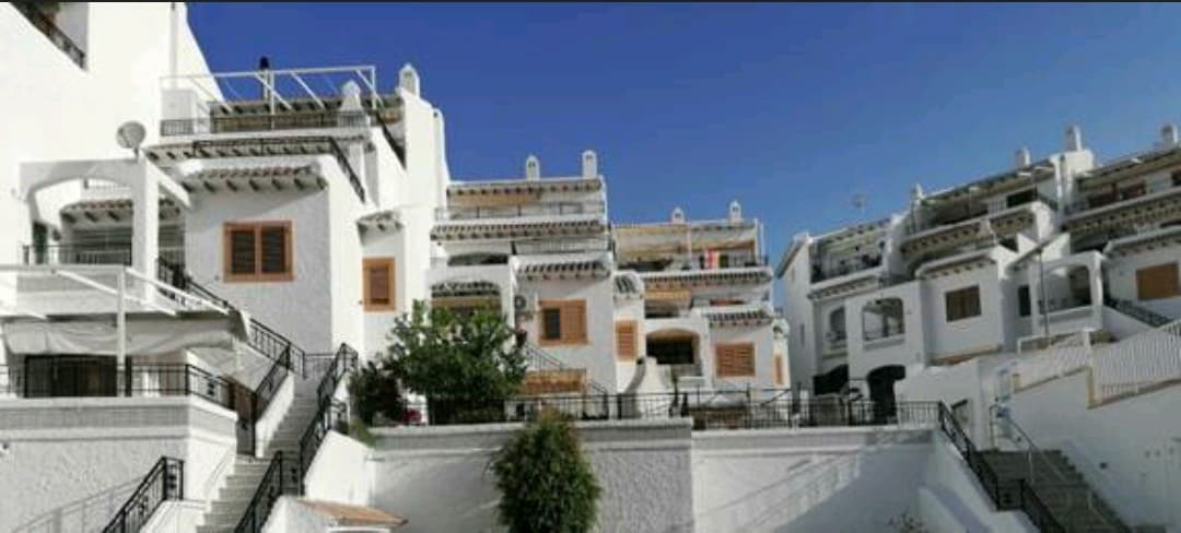 A series of well-maintained white apartment buildings are visible, arranged across a sloped landscape. Multiple terraces are adorned with potted plants, and staircases lead up between the buildings. The clear blue sky above enhances the atmosphere of the quiet residential community.
