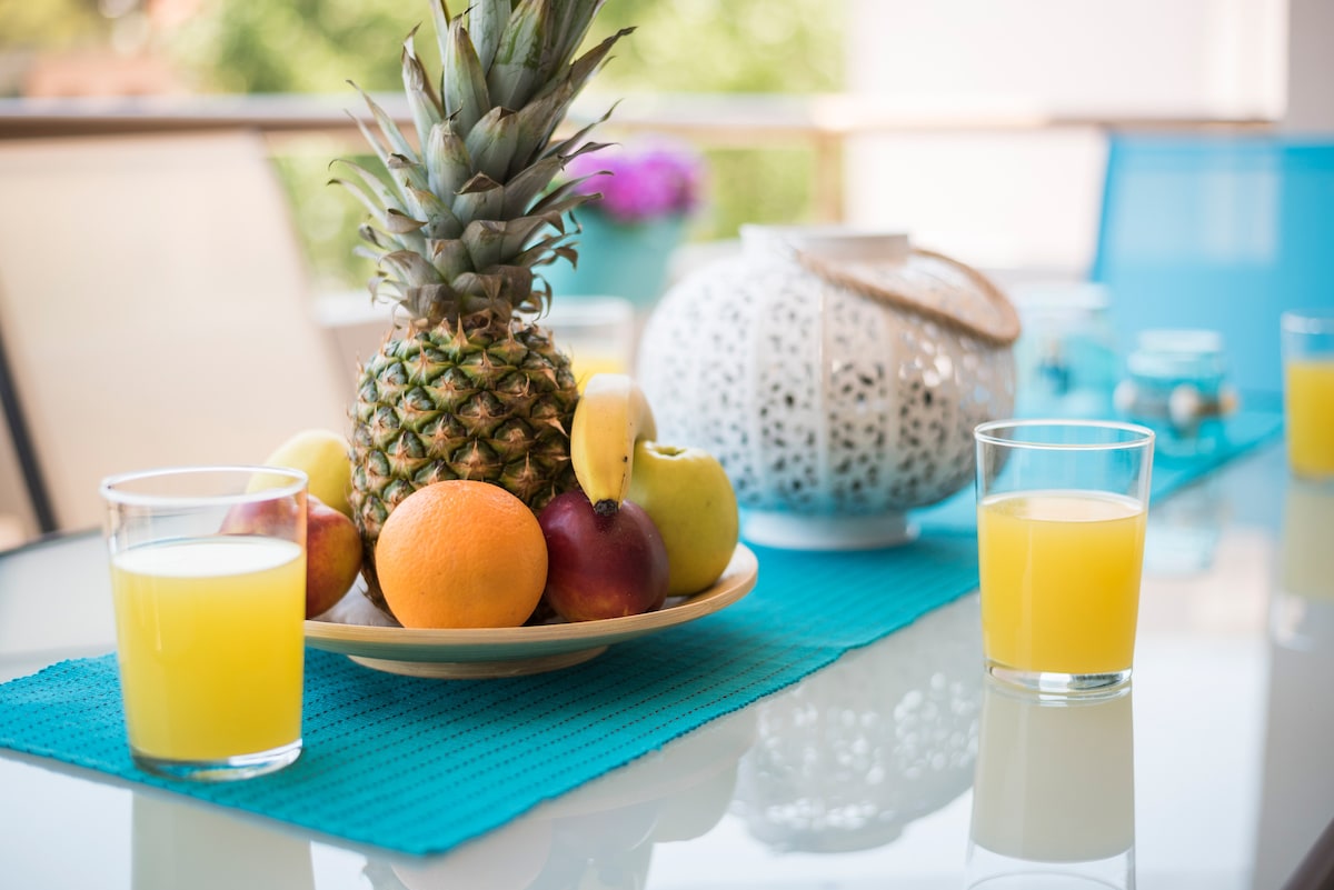 A vibrant table is set with a bowl of assorted fresh fruits, including a pineapple, apples, oranges, and bananas. Two glasses of yellow liquid, possibly juice, are placed on a turquoise tablecloth, enhancing the inviting atmosphere of the outdoor space.