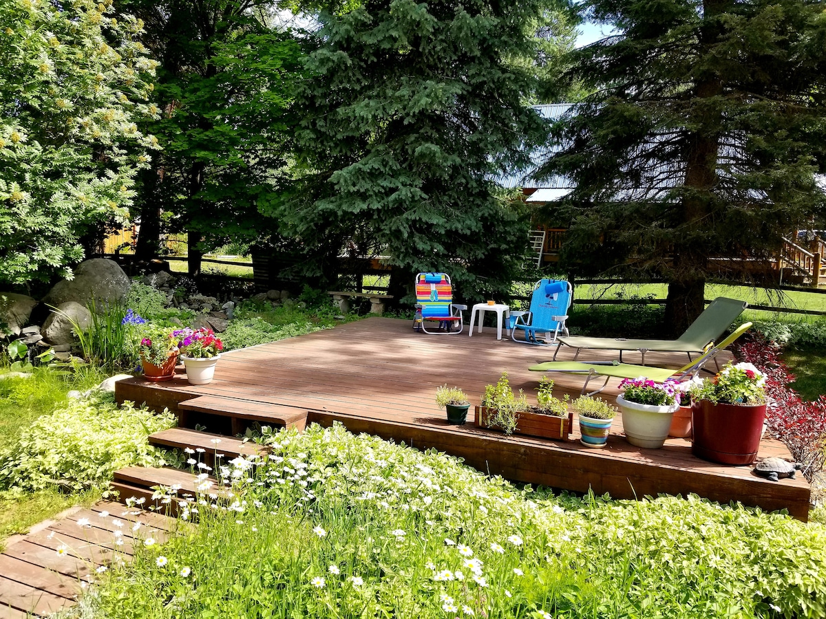 A spacious wooden deck is surrounded by vibrant greenery and flower pots, offering seating options including lounge chairs and small tables. The area is highlighted by a variety of colorful flowers and plants, providing a serene outdoor space for relaxation.