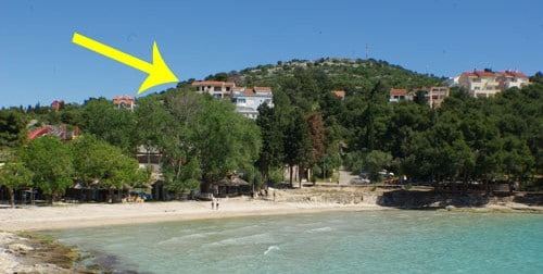The image shows a sandy beach with calm waters in the foreground, bordered by trees. In the background, an elevated hillside features residential buildings amidst greenery. The sunny sky highlights the vibrant landscape, providing a clear view of the peaceful beach area.