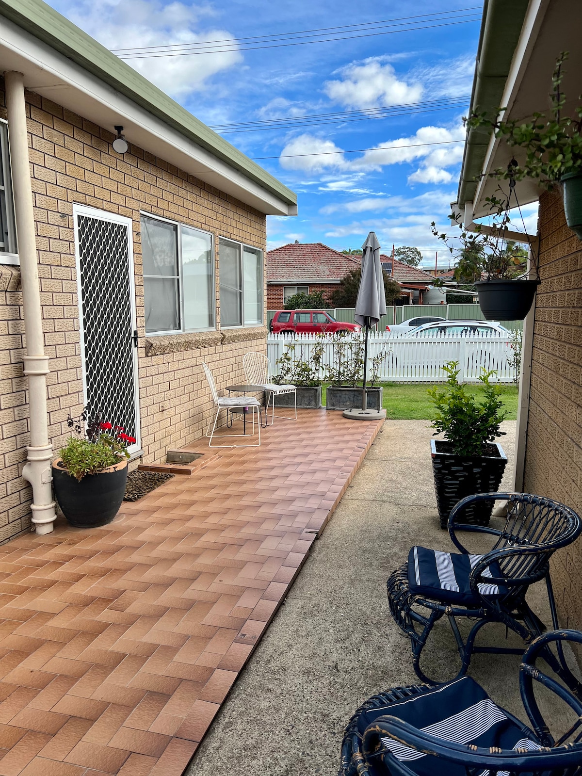 A well-maintained outdoor area features a paved path leading to a cozy seating corner with two chairs and a small table. Potted plants add greenery, while an umbrella provides shade. The scene is framed by attractive brick walls and a white picket fence in the background.