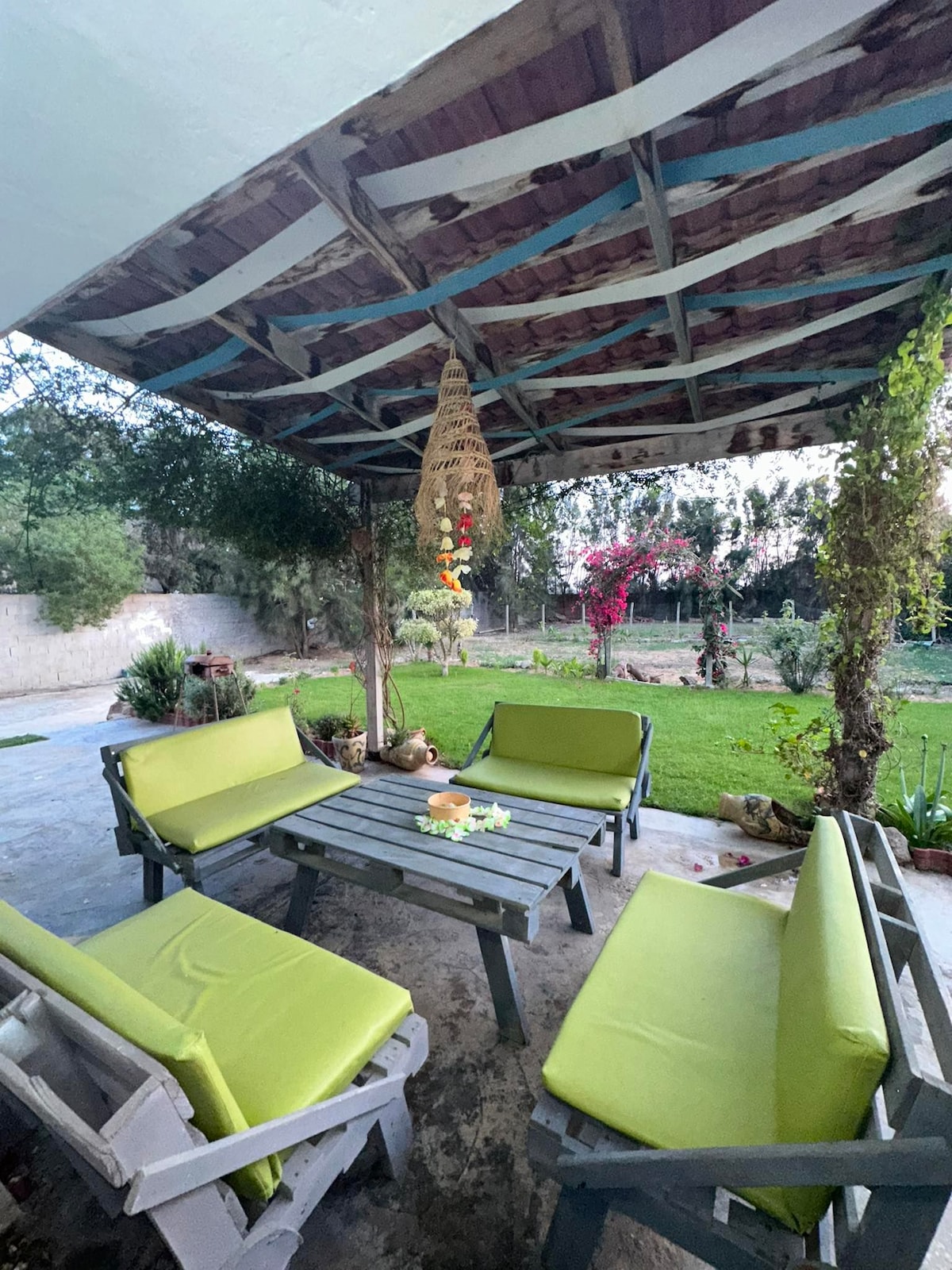 An outdoor seating area is featured with a wooden table surrounded by green cushioned benches. Above, a shaded canopy provides shelter, while a serene garden is visible in the background, showcasing lush greenery and flowering plants.