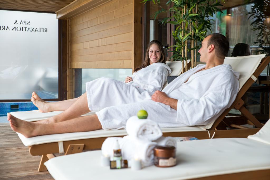 Two individuals are seated in spa robes on lounge chairs, positioned near a glass wall. Soft towels and spa products are arranged neatly on a nearby table. Lush greenery is visible, contributing to a calming atmosphere within the relaxation area.