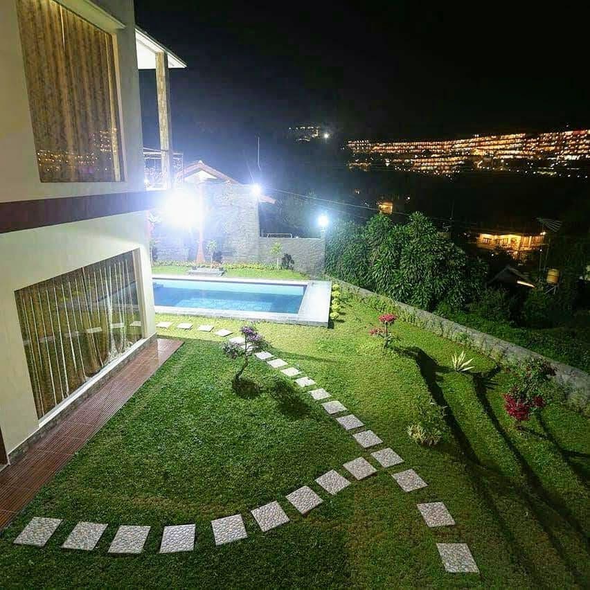 A well-lit garden area features a neatly maintained lawn and a rectangular swimming pool. Stone pathways lead through lush greenery, while vibrant flowers add color to the scene. The villa overlooks a distant illuminated landscape under the night sky.