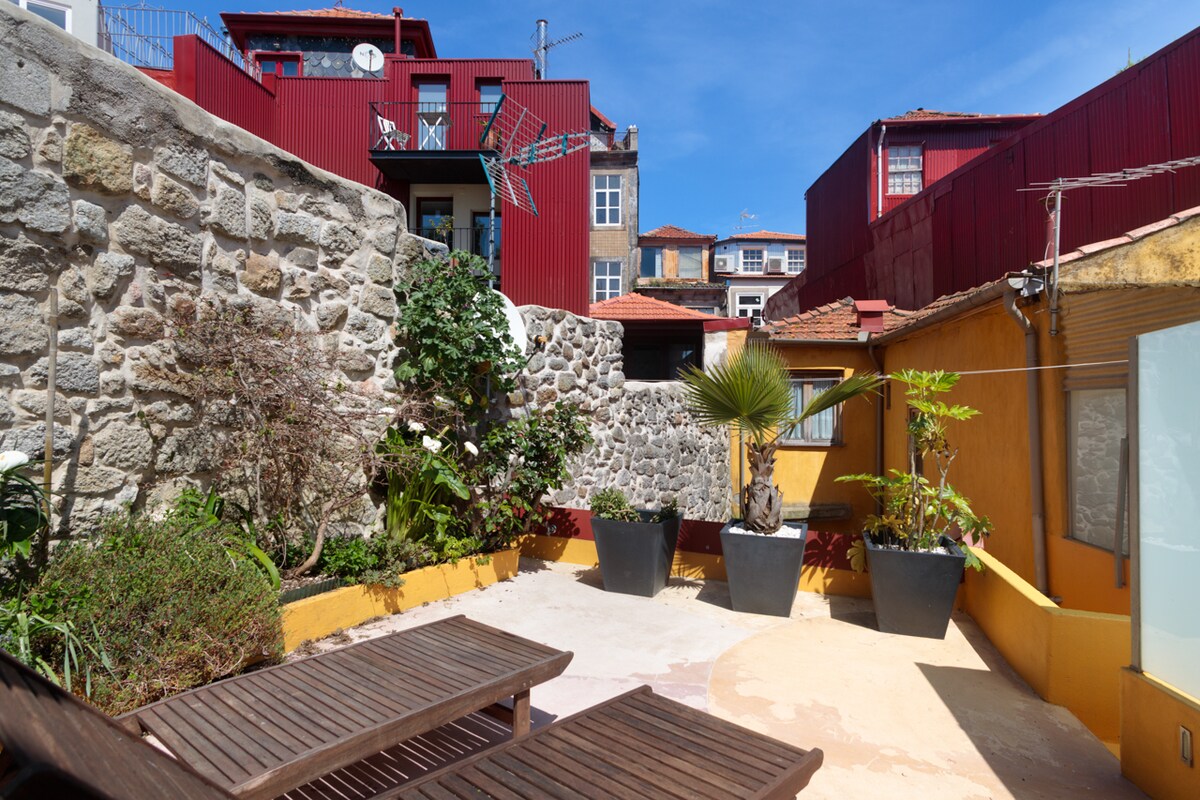 An outdoor garden space is showcased with two wooden lounge chairs positioned on sandy ground. Large planters filled with greenery are arranged among colorful walls, while a stone wall offers a rustic backdrop. The sky is clear, providing abundant sunlight.