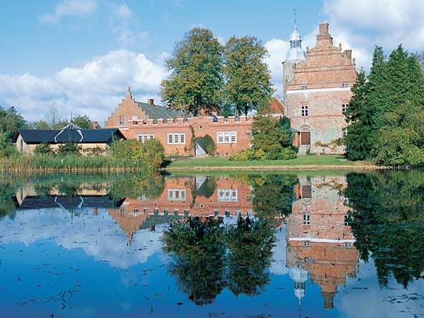 A historic building complex is reflected in a serene body of water, surrounded by lush greenery and trees. The structure features a mix of brick and stone elements, showcasing architectural details that suggest a long history. Soft clouds drift in the blue sky above.