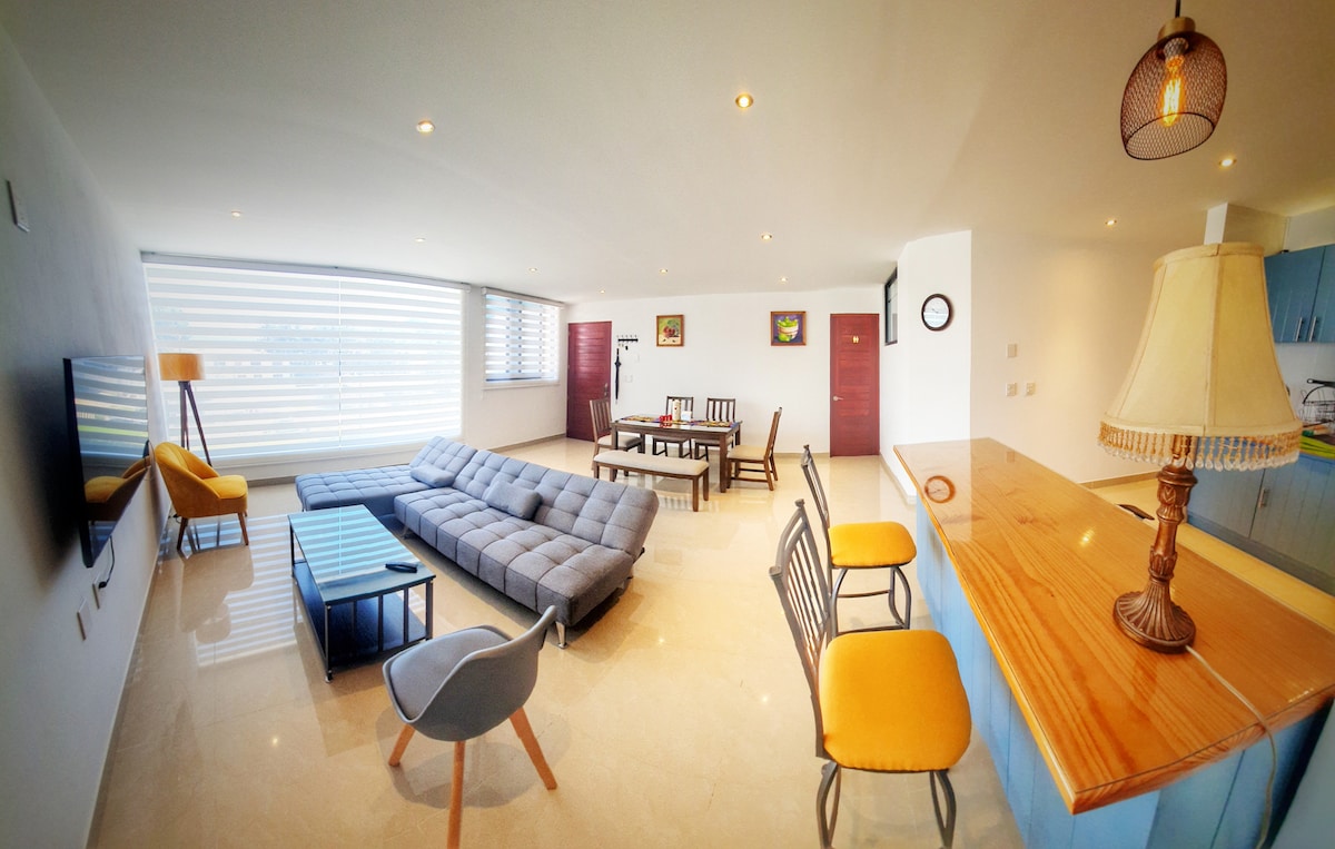 A spacious living area features a comfortable gray sectional couch arranged around a low coffee table. A dining table with chairs is positioned nearby, and light pours in through large windows adorned with sheer blinds. A kitchen bar with seating is visible in the background.