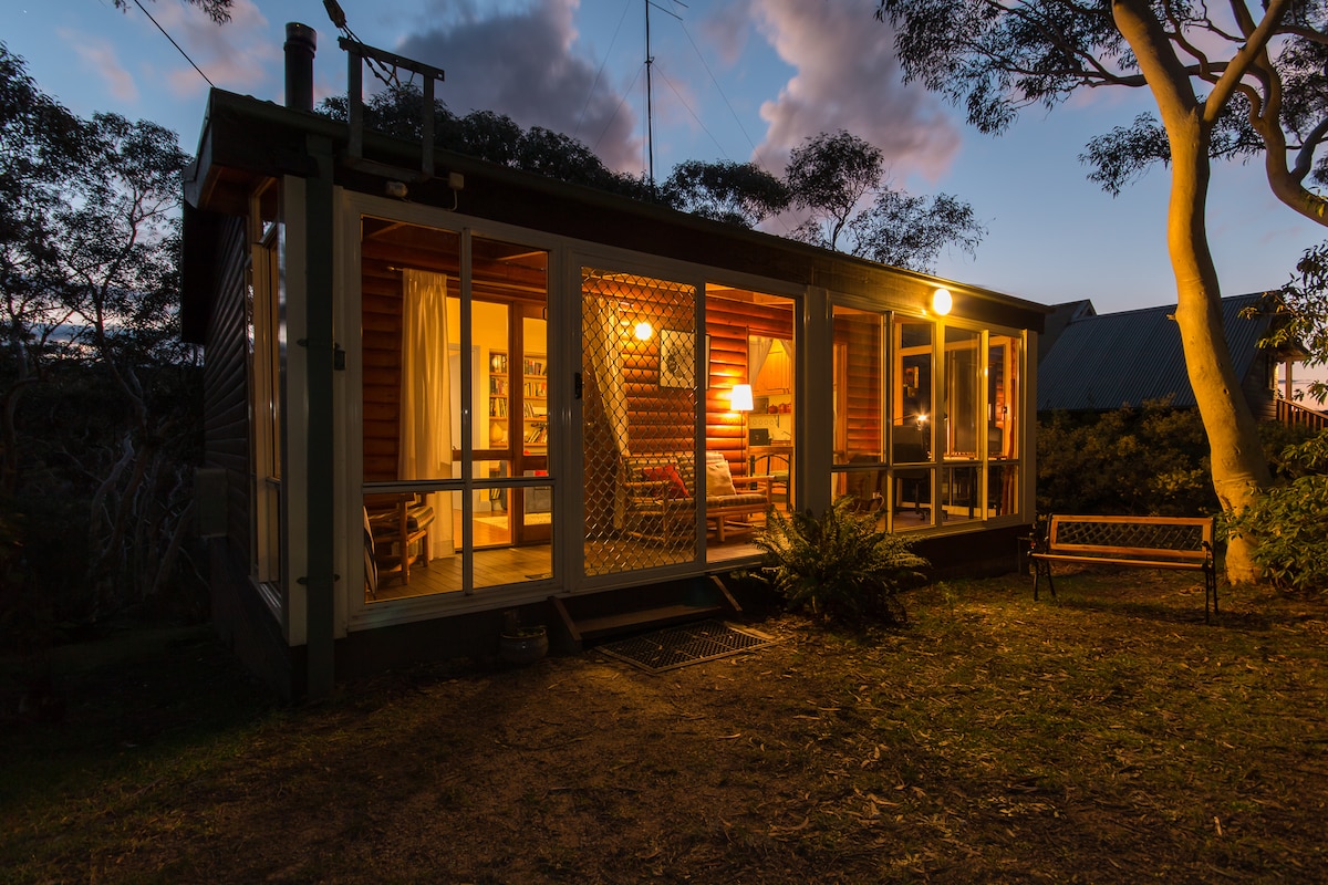 A log cabin-style cottage is seen at dusk, highlighted by warm light spilling from the windows. The structure features a spacious porch area with a comfortable seating arrangement. Surrounding bushland provides a natural ambiance, while a bench stands in the foreground, inviting relaxation.