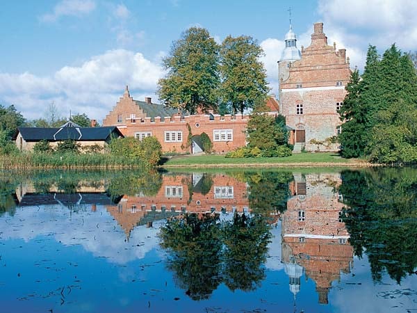 A historic building is reflected in a calm body of water, surrounded by trees and greenery. The structure features a combination of red brick and stone walls, topped with a tower. A pathway and additional buildings are visible nearby, enhancing the serene landscape.