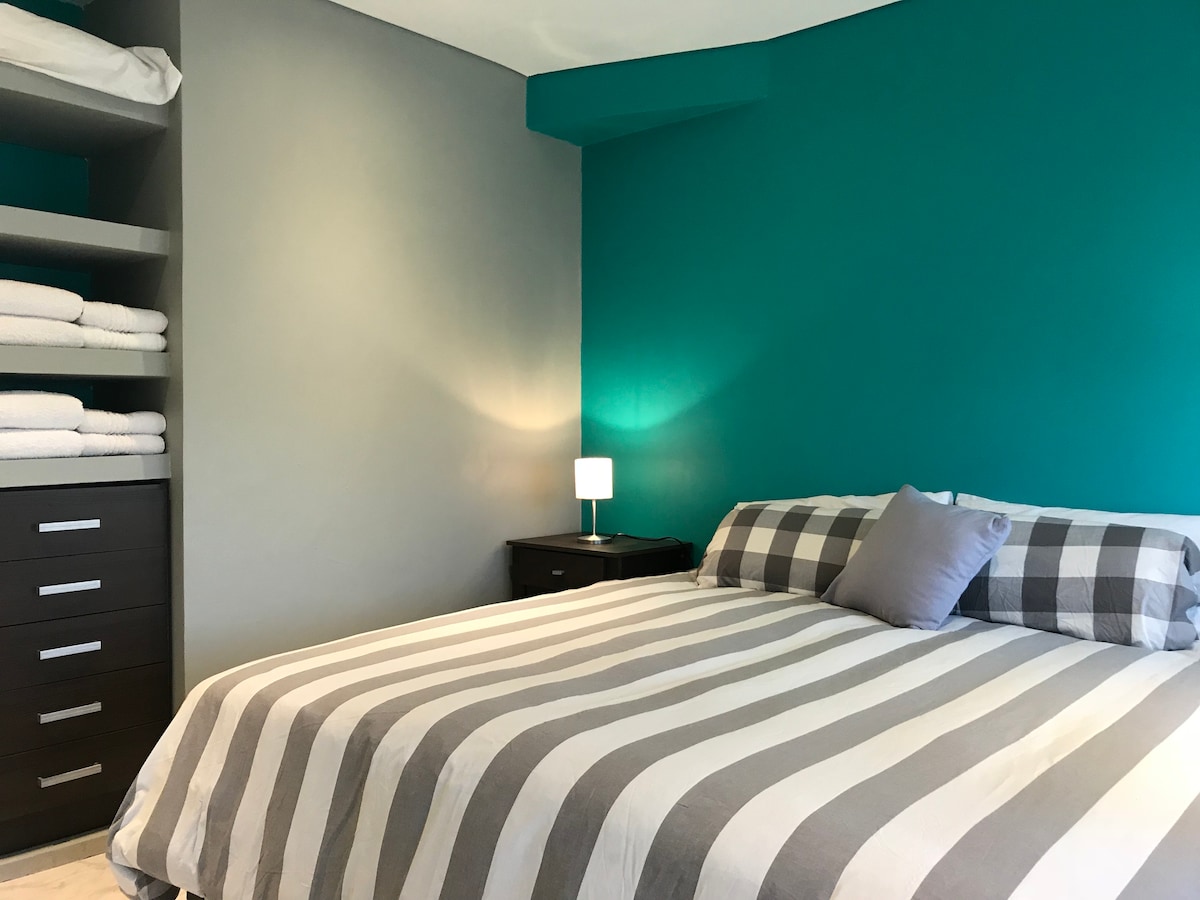 A spacious sleeping area is presented with a striped bedspread atop a large bed. Soft, neutral-colored pillows are arranged on the bed, and a small nightstand with a lamp is positioned nearby. Shelves with neatly stacked towels and a dresser enhance the functional design.