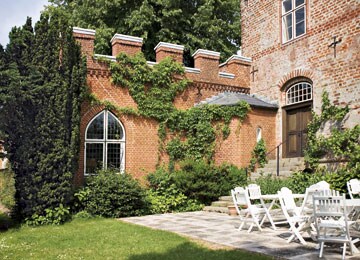 A charming brick exterior features an arched window surrounded by lush greenery. A small staircase leads to an entrance, while a stone patio is furnished with white outdoor chairs, inviting relaxation in the serene garden setting.