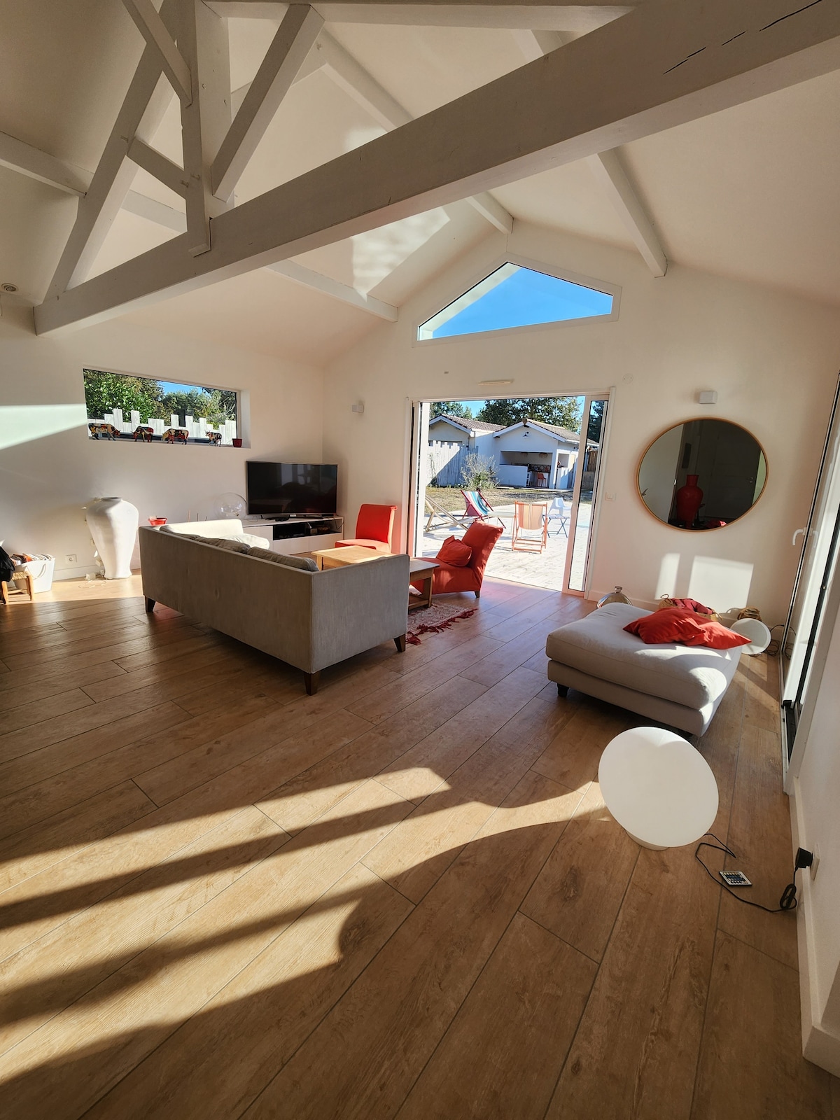 A bright living area features a high ceiling with exposed beams. A comfortable gray sofa and red accent chairs are arranged around a coffee table. Large glass doors lead to an outdoor space, allowing natural light to fill the room.