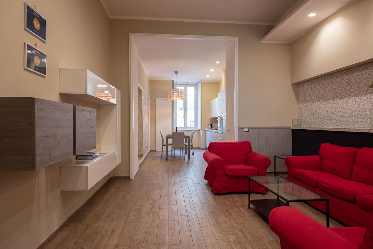 A welcoming living area is designed with red sofas surrounding a glass coffee table. A flat-screen TV is mounted on a modern wall unit. Natural light fills the space through a large window, and a dining table is visible in the background.