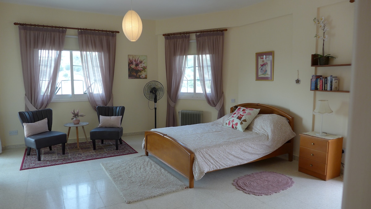 A spacious guest room features a large bed with a light bedding arrangement. Two armchairs with a small table are set in one corner. Natural light fills the room through two windows, framed with soft curtains, while a decorative fan adds comfort.