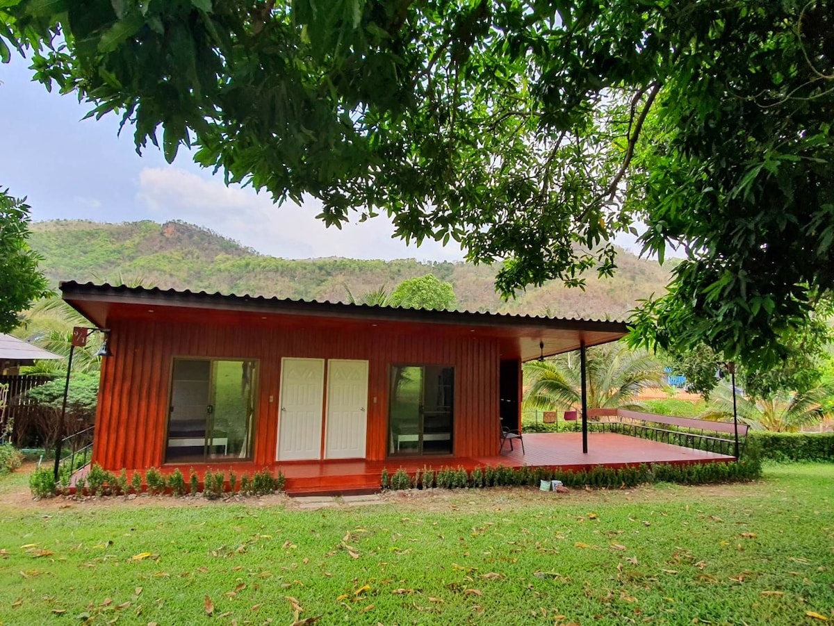 A modern house features a red exterior with large glass doors that provide a view of the surrounding greenery. The spacious terrace is accessible from the house, complemented by a serene backdrop of hills and a well-manicured lawn.