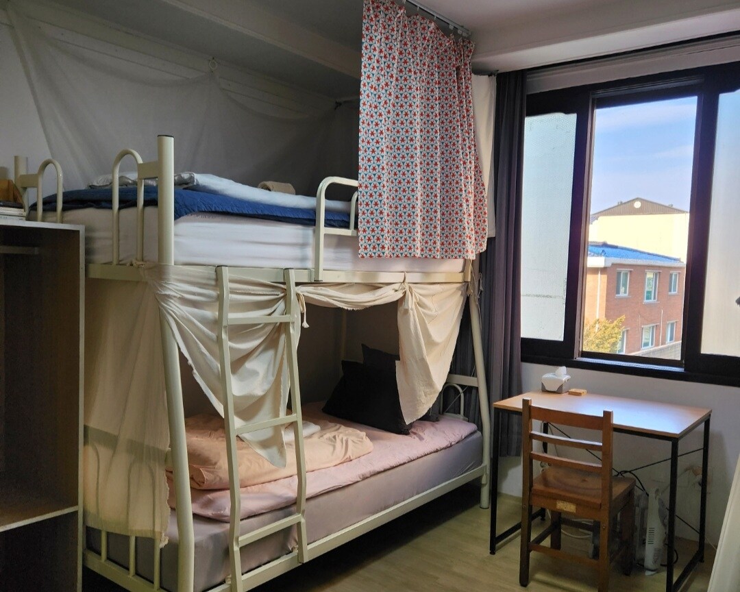 A cozy mixed dorm room features a bunk bed with a lower sleeping area draped in soft fabric. A simple wooden table and chair are positioned near a window, allowing natural light to enter. Light-colored walls and flooring enhance the room's inviting ambiance.
