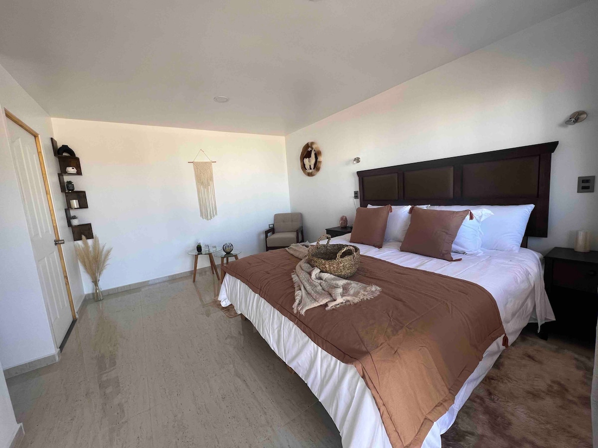 A spacious room is shown, featuring a large king bed with soft, neutral-toned bedding. A cozy armchair is positioned nearby, with decorative accents on the walls. Natural light fills the space, highlighting the minimalist decor and maintaining an inviting ambiance.