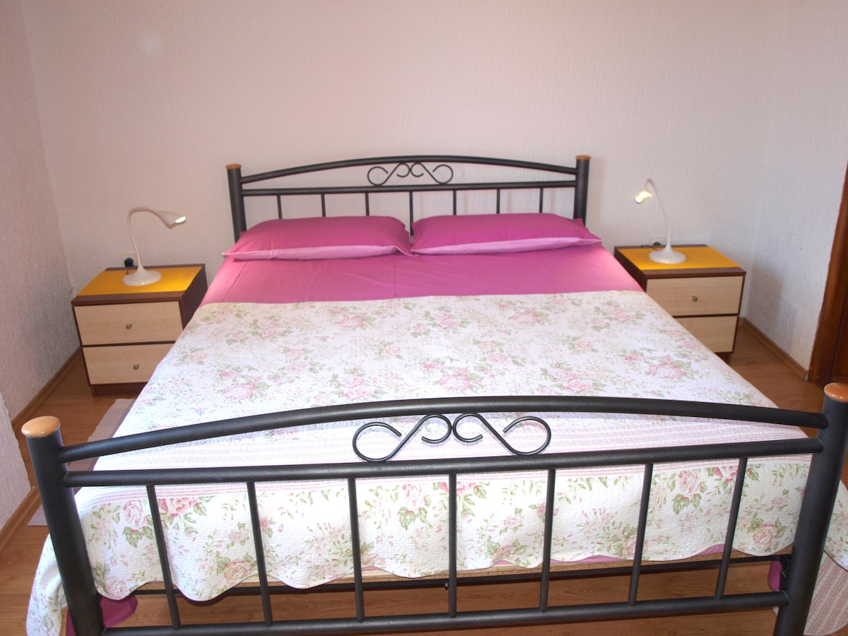 A spacious double bed is dressed with floral-patterned bedding, surrounded by two bedside tables with lamps. Soft purple linens add a touch of color, while wooden flooring contributes to a warm ambiance in the bedroom.