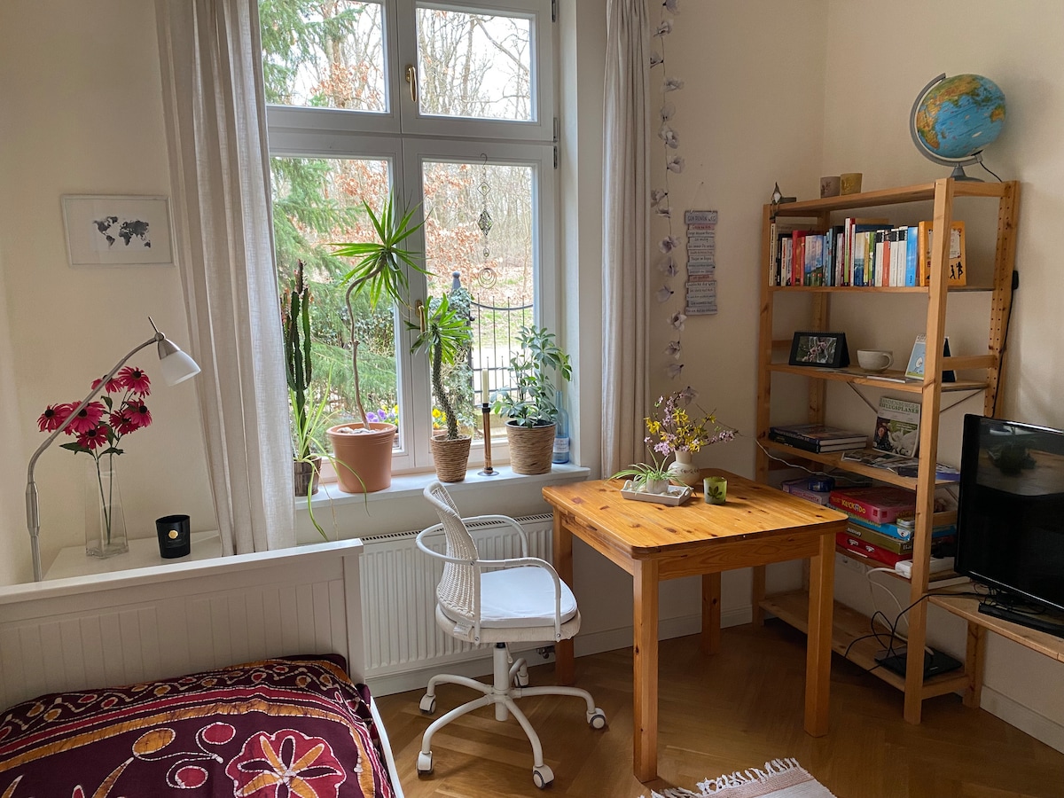 A bright room features a window with a view of greenery, adorned with potted plants. A wooden table with a chair is positioned beside the window. A small bookshelf holds various items, while a cozy bed is partially visible, covered with a patterned blanket.