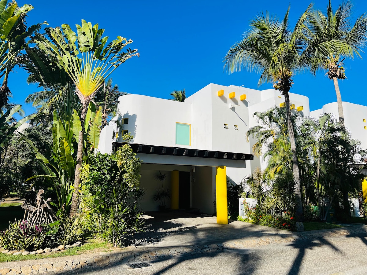 A modern villa is set amidst lush tropical plants and palm trees. The entrance features a covered drive leading to a white facade accented by vibrant yellow details. The clear blue sky provides a bright backdrop, enhancing the property's inviting appearance.