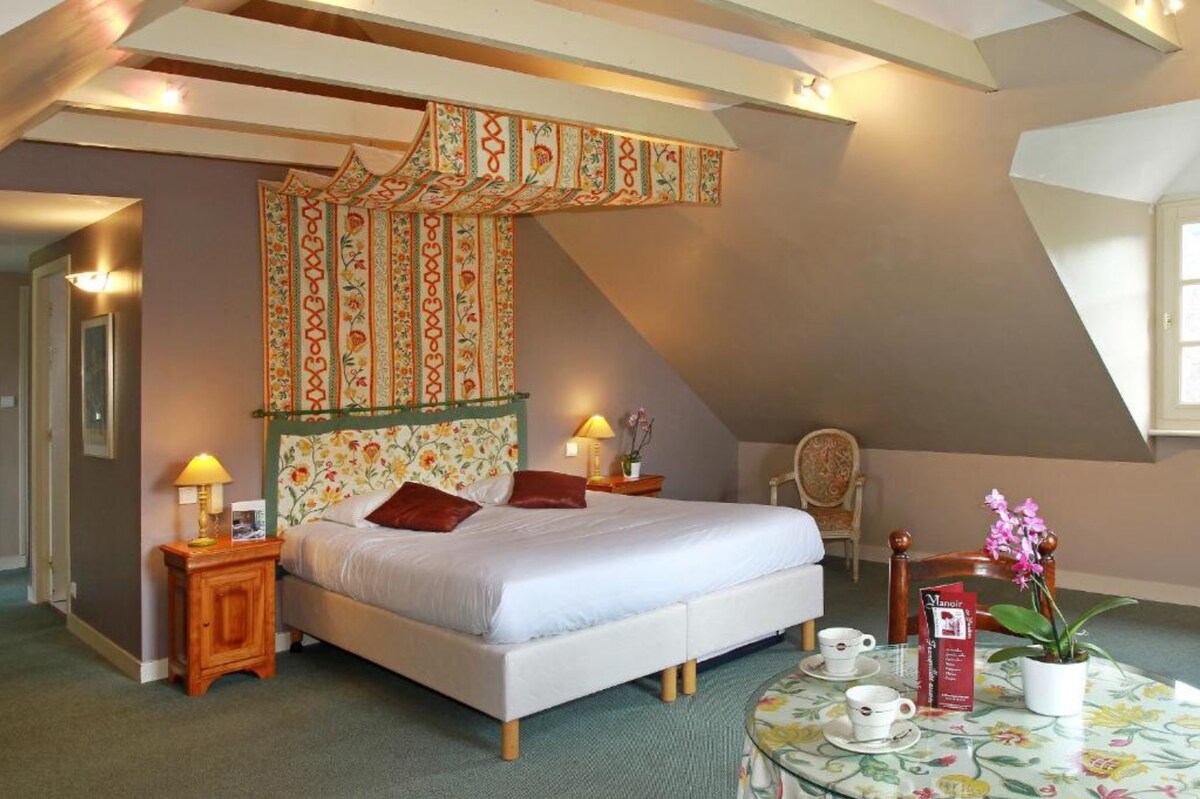 A spacious room features a large bed with patterned headboard and neutral bedding. Two lamps provide soft lighting. A small seating area includes an elegant chair beside a flower arrangement. A dining table is set with delicate tableware, enhancing the inviting ambiance.